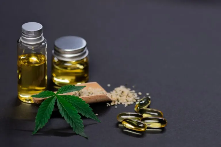 Glass jars of cannabis oil with a hemp leaf, hemp seeds and softgel capsules on a dark surface